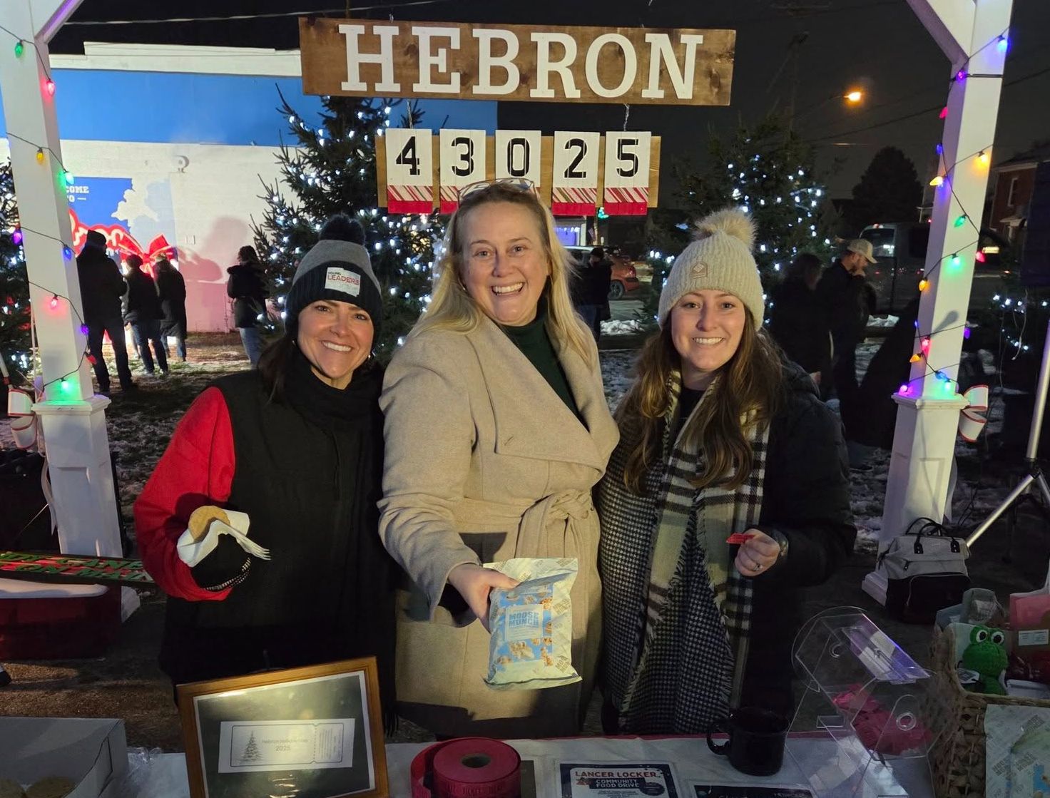 Three women stand smiling behind a table at a festive Hebron community event.