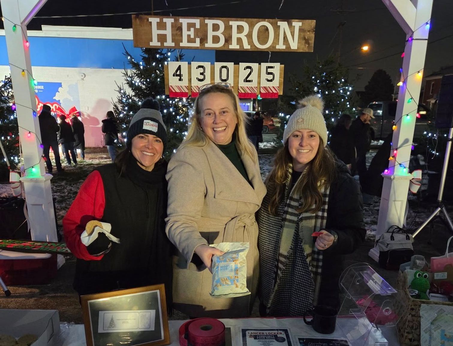 Three women stand behind a table at a night market under a 'HEBRON EGGIE' sign.