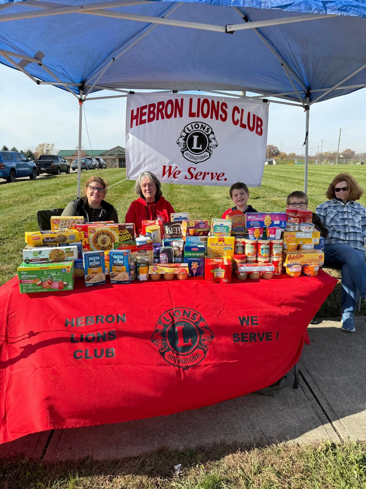 Hebron Lions Club volunteers stand behind a table full of donated food items under a blue canopy.