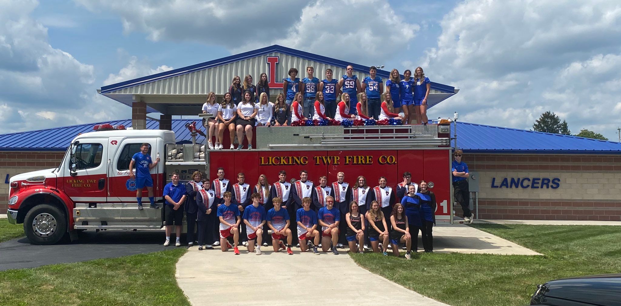Band members in uniform pose with a fire truck in front of a school building.
