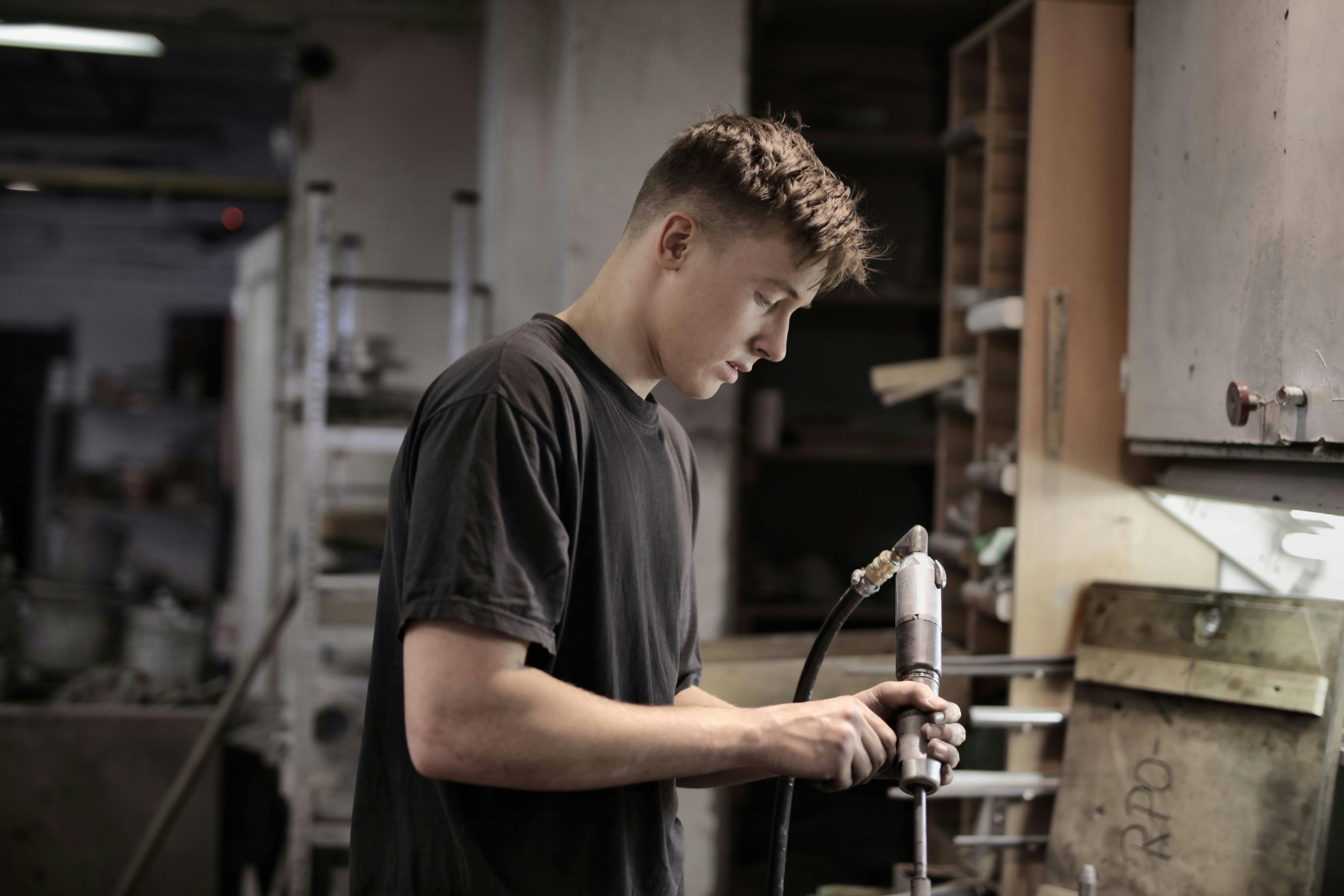 Young man with short brown hair intently operates a mechanical tool in a dimly lit workshop.