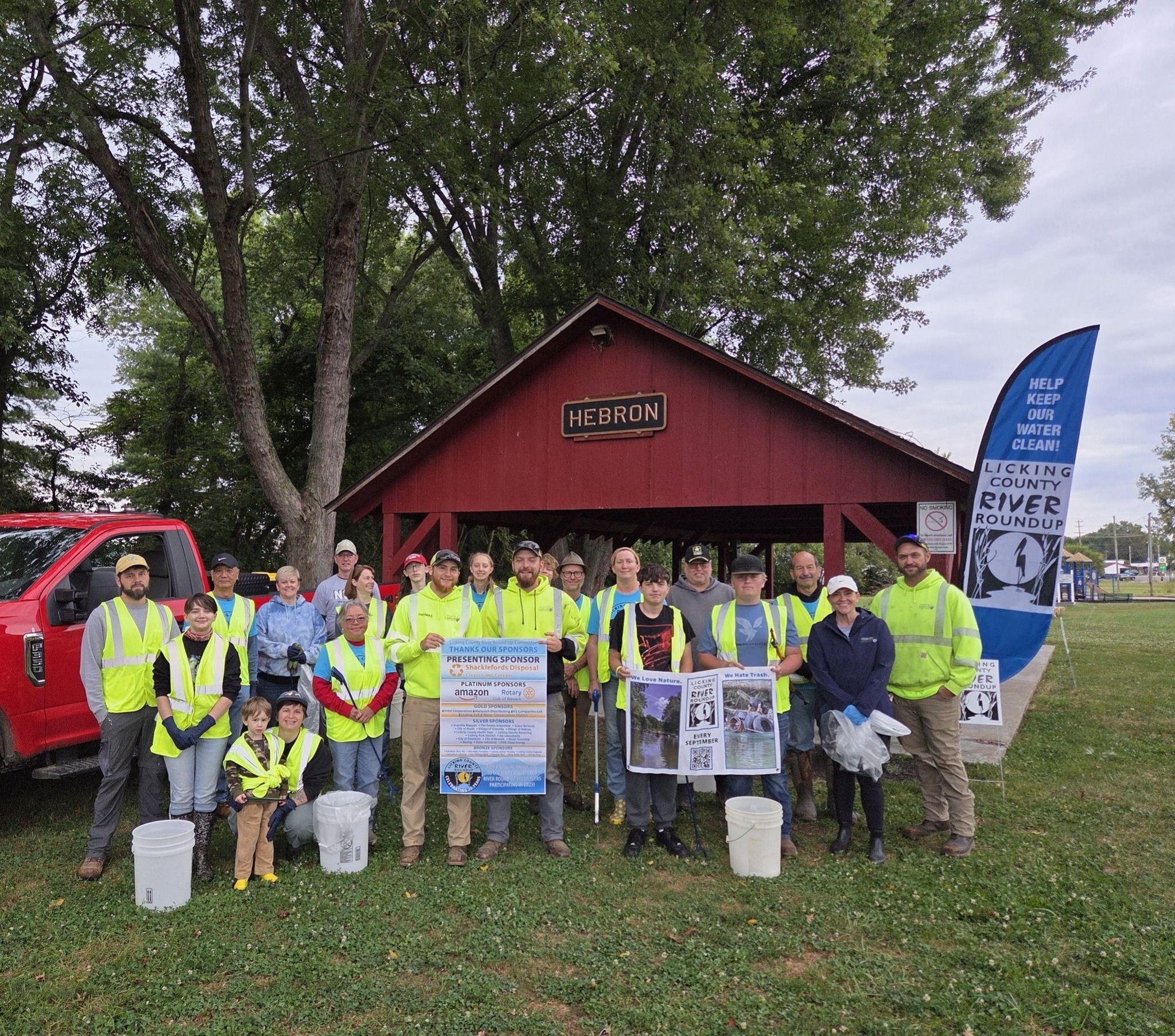Volunteers in high-visibility vests stand with cleanup buckets and signs in front of a red building.